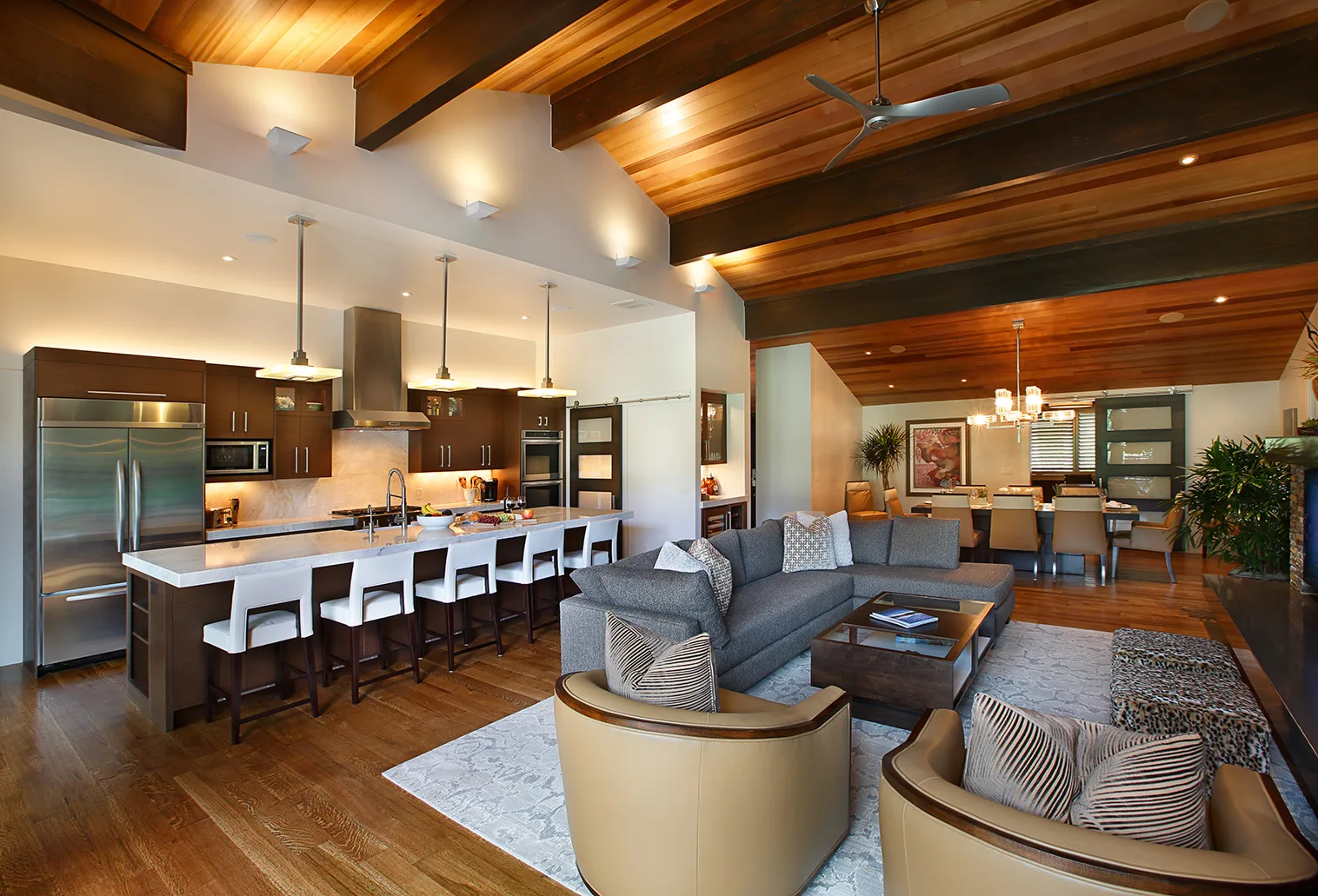 Open-plan kitchen with a long island and bar stools opening to a living area with a round ottoman and warm wood tones