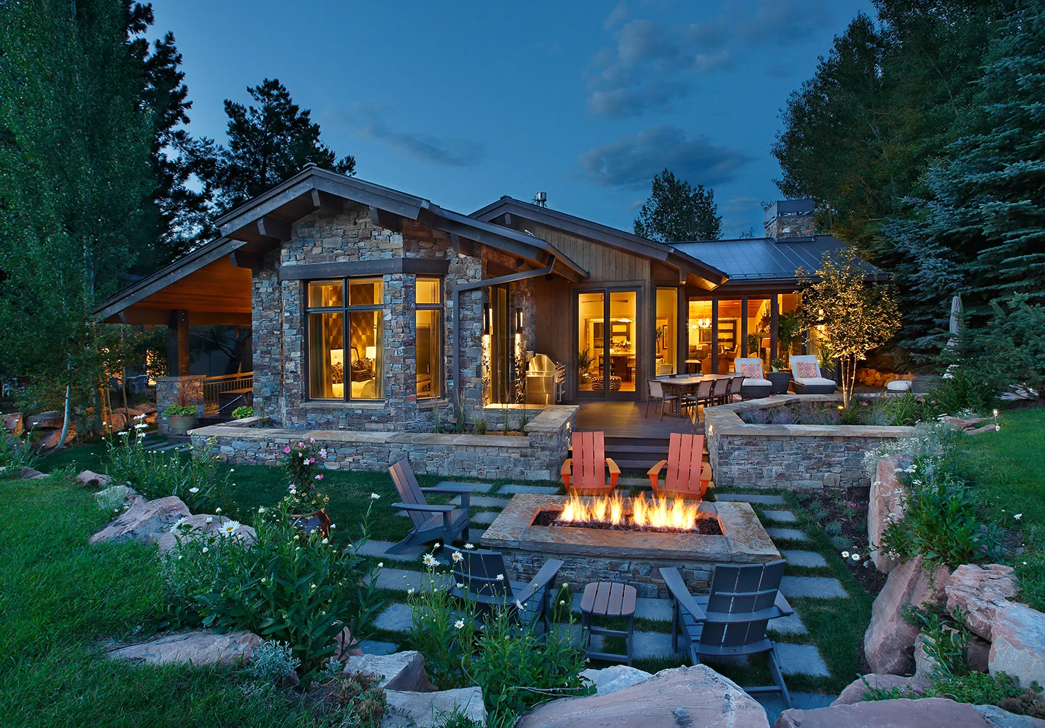 Dusk exterior of a stone and timber mountain home with a lit fire pit, seating area and snow-capped peaks behind